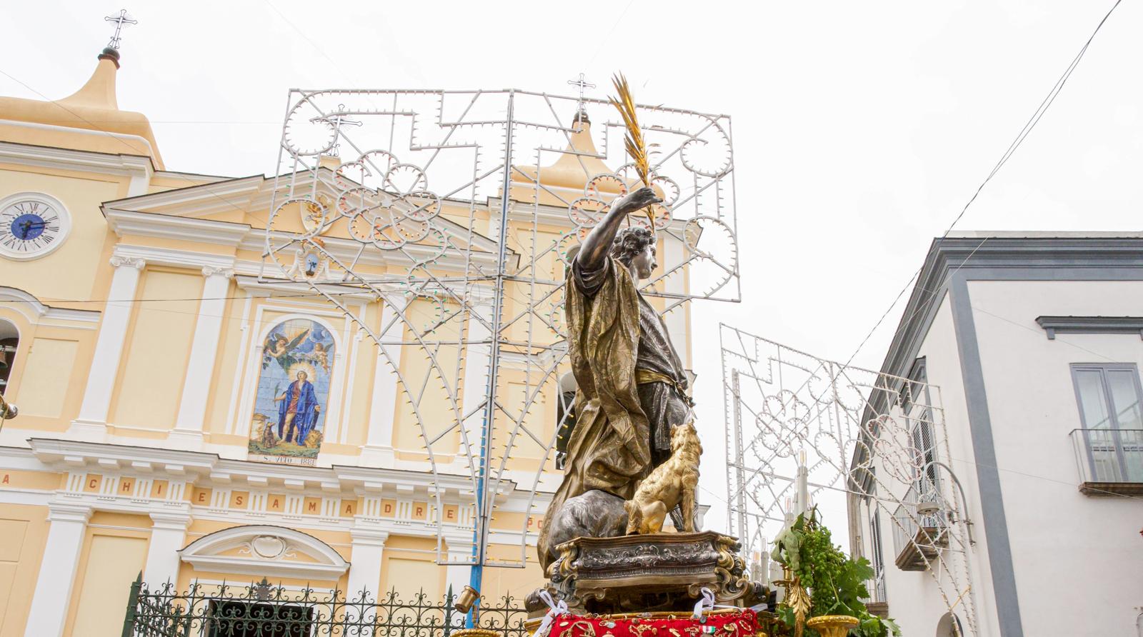 Il santo patrono, in primo piano, è raffigurato in una statua che lo ritrae mentre tiene una palma nella mano destra, simbolo di vittoria e martirio. Alle sue spalle si erge la Basilica, la chiesa madre del patrono, con la sua facciata imponente che domina la scena. Sullo sfondo, le luminarie che decorano la piazza creano una suggestiva atmosfera di festa, rendendo ancora più luminosa e vivace la celebrazione patronale.
