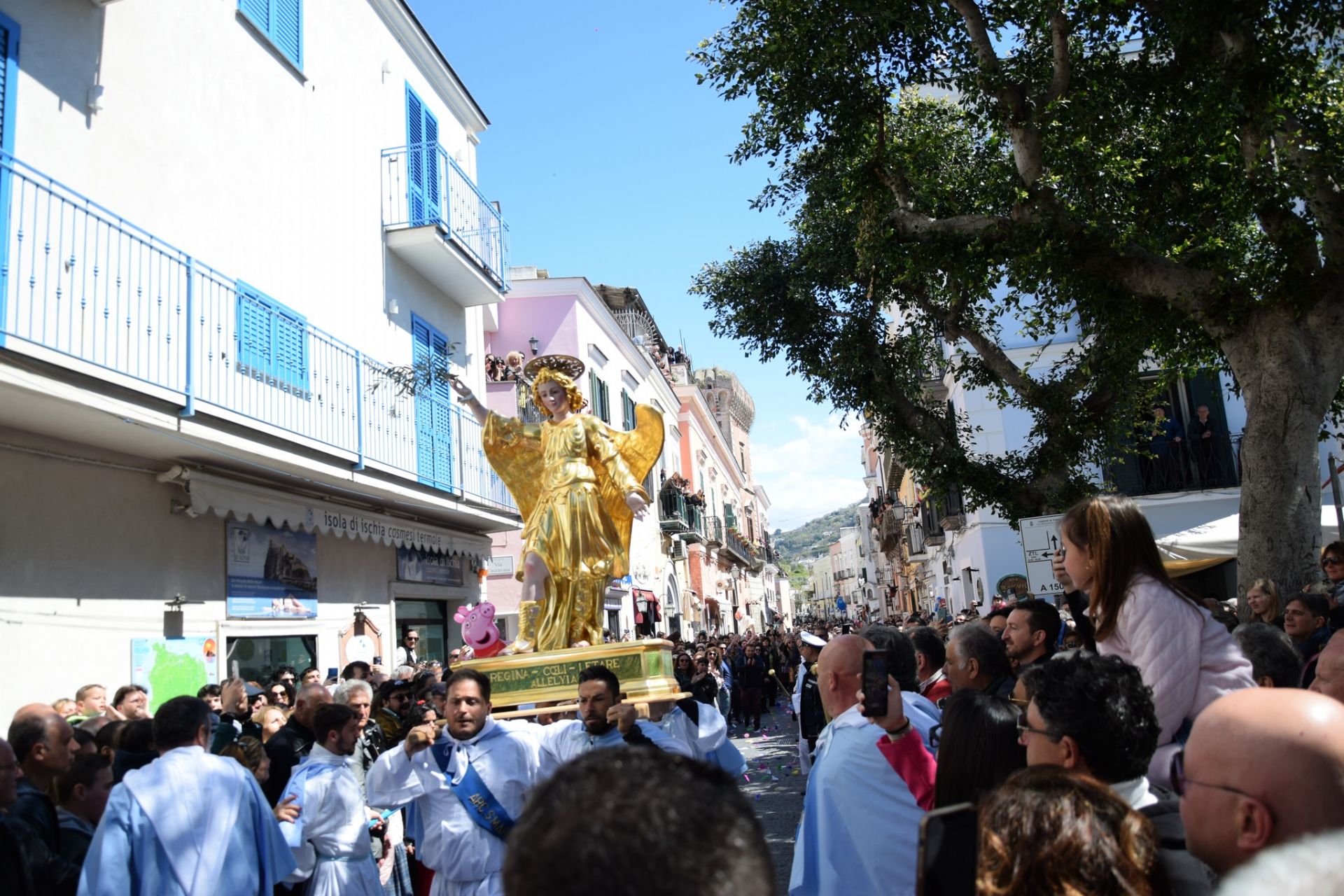 La foto mostra un momento della tradizionale Corsa dell'Angelo a Forio, con un angelo in volo, circondato da una folla entusiasta che celebra l'evento religioso e culturale.