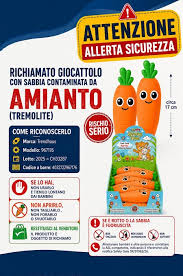 giocattolo a forma di carota sorridente, arancione con ciuffo verde. Scritta di allarme Richiamo giocattolo con sabbia contaminata da amianto.